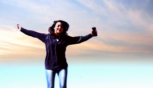 Young man with arms raised standing against sky during sunset