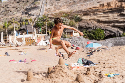 Young man wearing a swimsuit jumping over a sand castle at exotic beach near the sea. summer concept