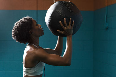 Side view of woman holding medicine ball while listening music against wall in gym