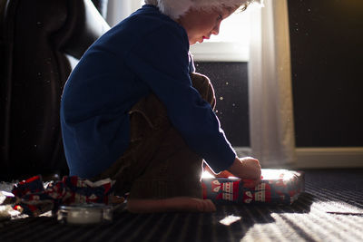 Boy in warm clothing wrapping present while crouching on carpet at home