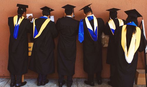 Rear view of students in mortarboards writing on paper against wall