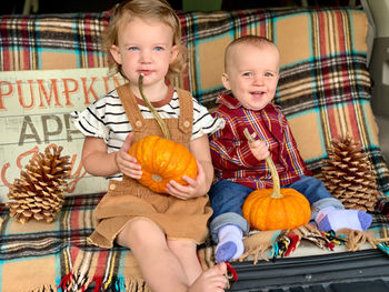 Cute siblings with pumpkins and pine cones sitting on bench