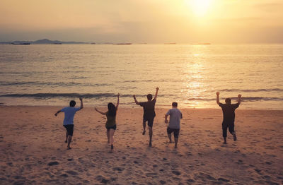 Rear view of friends jumping at beach against sky during sunset