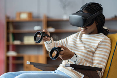 Happy young african american woman wearing virtual reality goggles playing video games at home