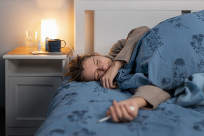 Young woman holding thermometer resting on bed at home