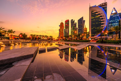 Reflection of illuminated buildings in lake against sky during sunset