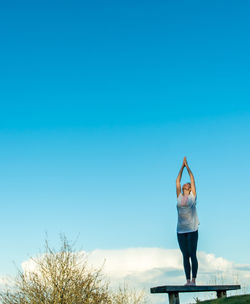 Full length of woman doing yoga on bench at park against blue sky