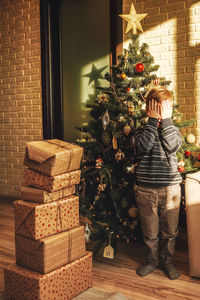 Boy with hands covering eyes standing by christmas tree and gifts at home