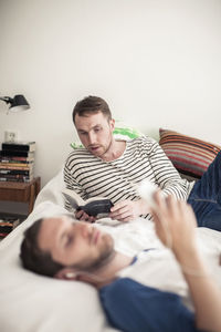 Young gay man reading book while partner listening music in foreground on bed at home
