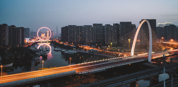 Illuminated bridge over river amidst buildings in city against sky