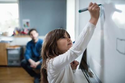 Confident girl drawing on blackboard while teacher sitting in background