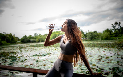 Young woman drinking glass at water against sky