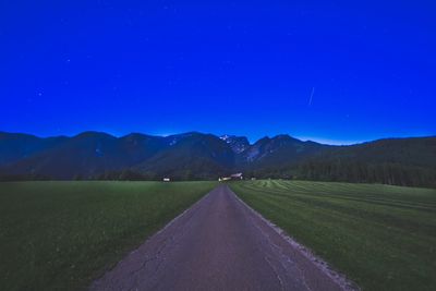 Empty road amidst field and mountains against blue sky