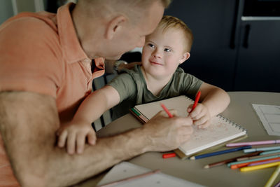 Boy with down syndrome looking at father while drawing on book