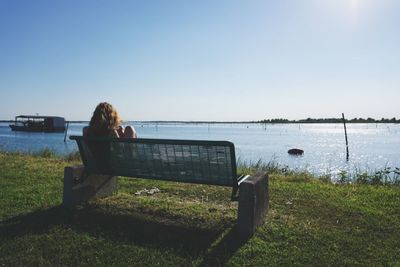 Woman sitting on grass by sea against clear sky