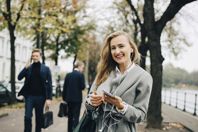 Smiling businesswoman with in-ear headphones and smart phone standing in city