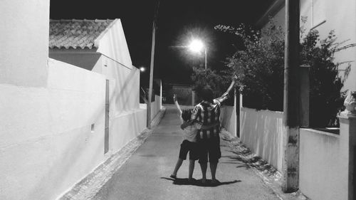 Rear view of boy and son with arms raised standing on footpath amidst houses at night