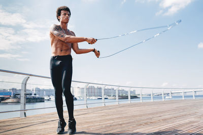 Shirtless young man jumping rope on promenade against sky