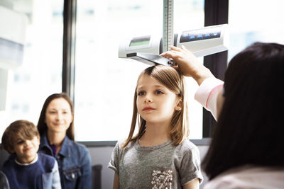 Doctor measuring girl's height while family sitting in clinic