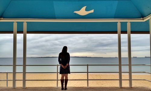 Rear view of woman standing in gazebo at beach against cloudy sky