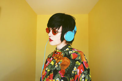 Young woman wearing blue headphones standing against yellow wall