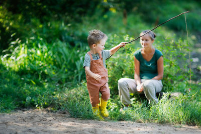 Mom and son in park among grass are playing fishing with stick and fishing rod. 