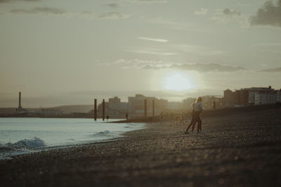 Man walking on beach in city against sky during sunset