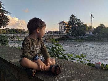 Boy looking at river while sitting on retaining wall against sky