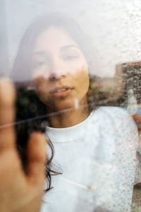 Through glass of young ethnic female gently touching window with raindrops and looking at camera