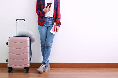 Low section of woman standing by luggage on hardwood floor against wall