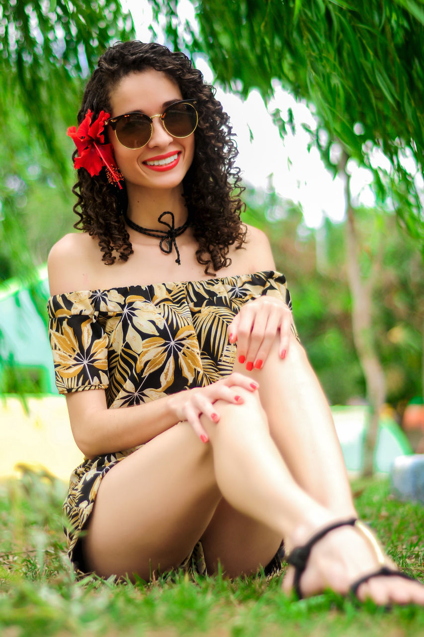 sunglasses, one person, beautiful woman, young adult, young women, outdoors, real people, leisure activity, fashion, lifestyles, day, front view, smiling, looking at camera, nature, happiness, portrait, grass, curly hair, tree