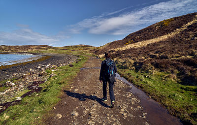 Rear view of woman walking on dirt road by mountain and sea against sky