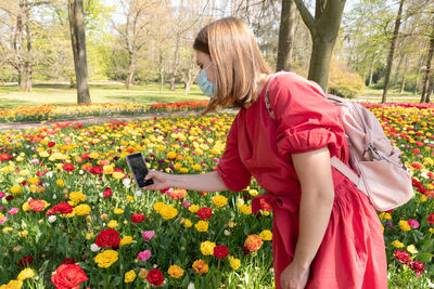 A blonde girl in a red dress and in a protective mask is photographing a flowers