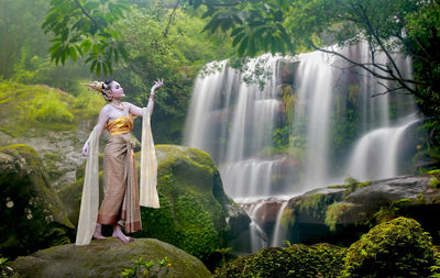 Full length of young woman in traditional clothing standing on rock against waterfall