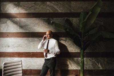Smiling senior businessman holding coffee cup talking through mobile phone leaning on wall at office