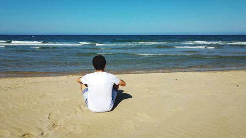 Rear view of man sitting on sand at beach against clear blue sky