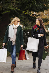 Smiling women with shopping bags looking at each other walking on footpath