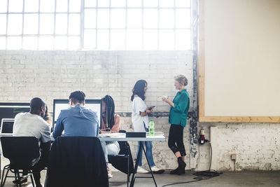 It experts coding computer programs while businesswomen talking by wall at creative workplace