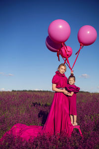 Mother and daughter in pink dresses are standing on a field with flowers at sunset in summer