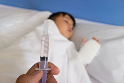 Cropped hand of person holding syringe against boy sleeping on bed at hospital
