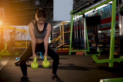 Full length of young woman lifting kettlebells while standing in gym