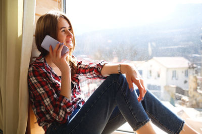 Young woman using mobile phone while sitting on window