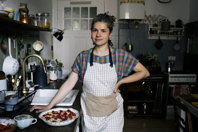 Portrait of woman with hand on hip standing by pie on kitchen counter at home