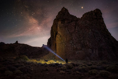 Silhouette person holding illuminated flash light by mountain at night