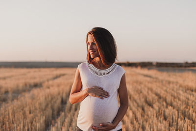 Smiling pregnant woman with hands on stomach standing on field