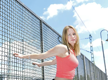 Portrait of young woman leaning on fence against cloudy sky