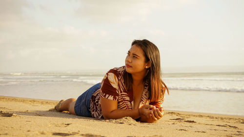 Young woman sitting on shore at beach against sky