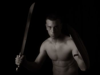 Portrait of shirtless teenage boy holding sword and shield against black background