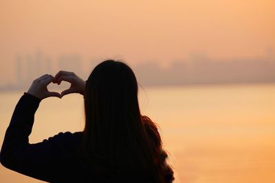 Rear view of woman making heart shape against sea during sunset