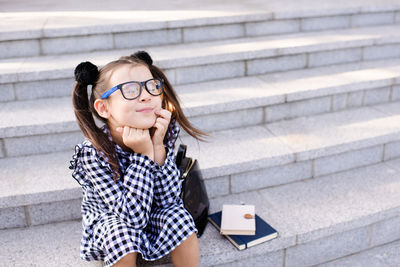 Funny pretty kid girl wear glasses and stylish dress sitting on stairs with books, backpack outdoors